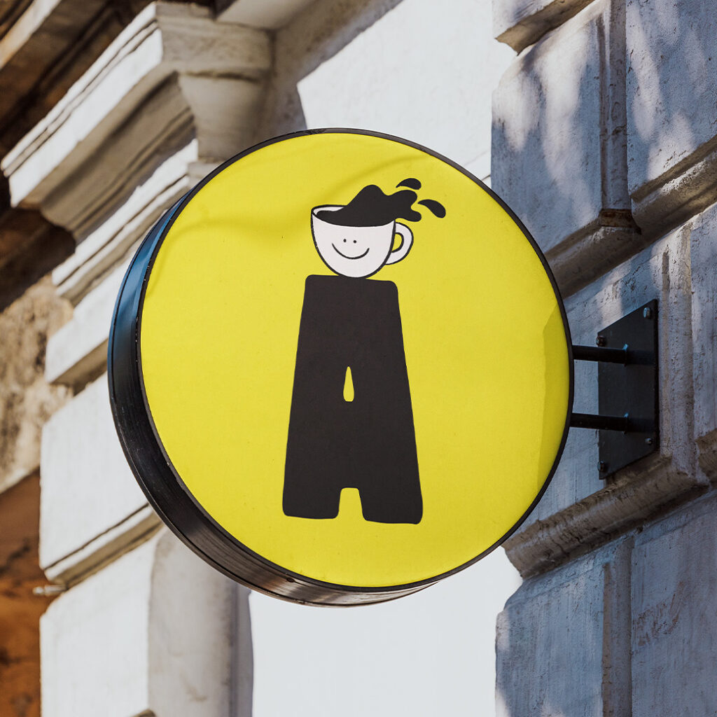 Enseigne extérieure ronde jaune avec le logo d’Agonista : une tasse de café souriante sur un pilier noir en forme de lettre A